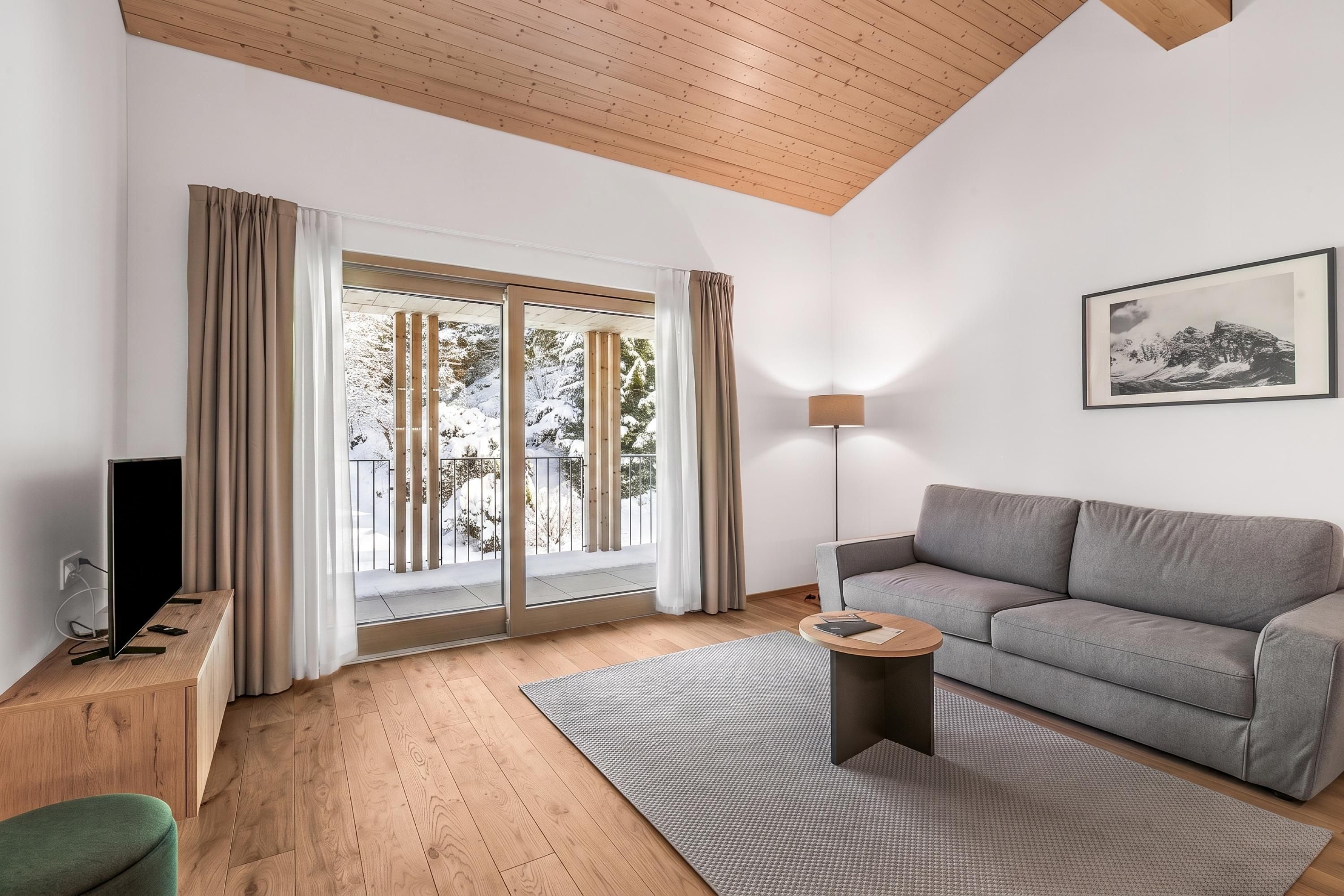A modern living room with a grey sofa, a flat-screen TV, a wooden floor, and a large window with curtains.