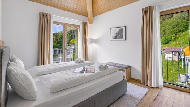 Ein modernes Schlafzimmer mit einem großen Bett, einem Nachttisch, einer Lampe und Blick auf eine grüne Landschaft durch das Fenster.