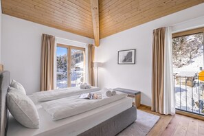 Ein Hotelzimmer mit Bett, Nachttisch, einem Fenster mit Vorhängen, einem Bild an der Wand und Blick auf eine verschneite Landschaft.