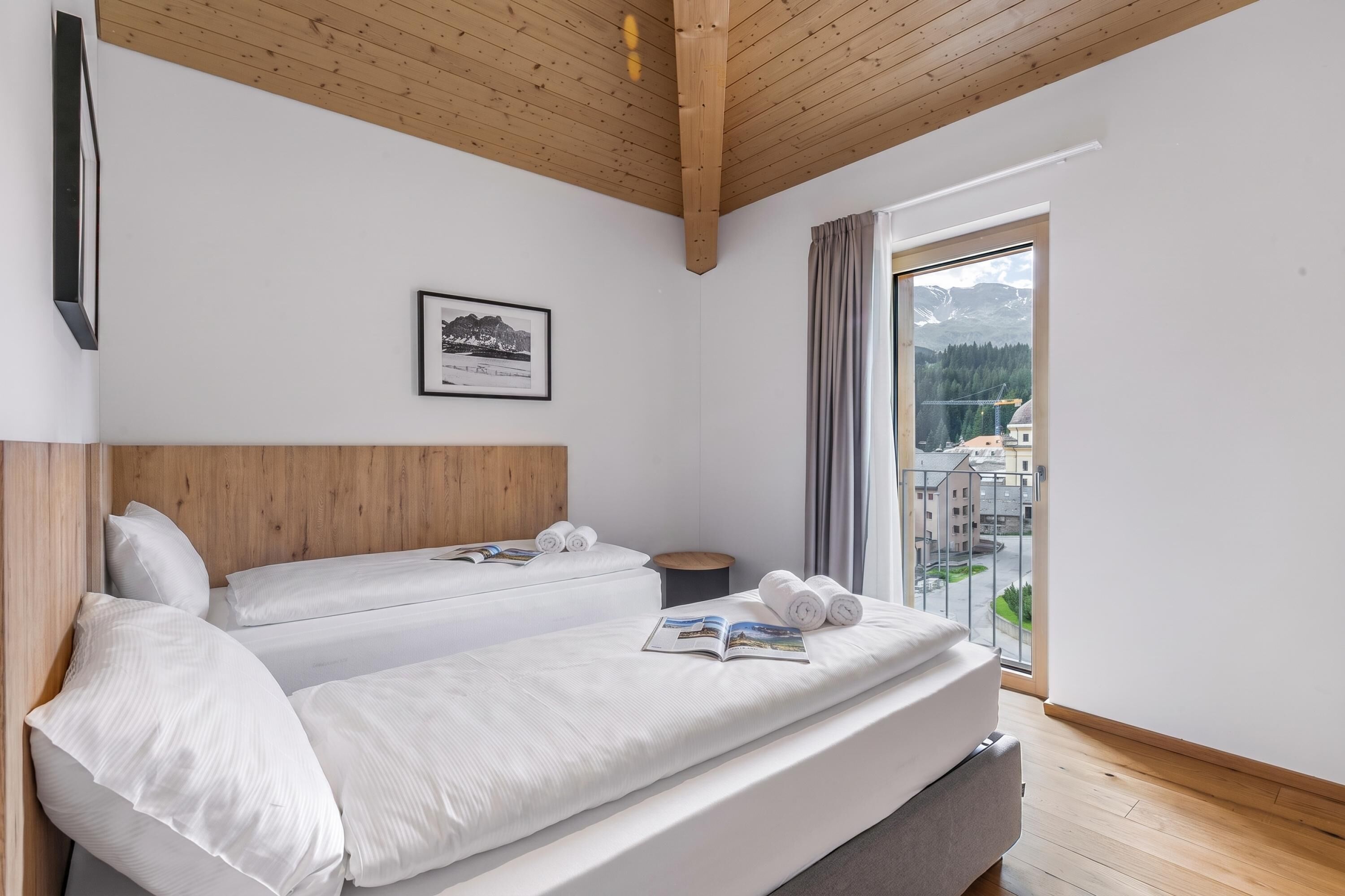 Habitación de hotel con dos camas, techo de madera, un cuadro en la pared y ventana con vistas a edificios y montañas.