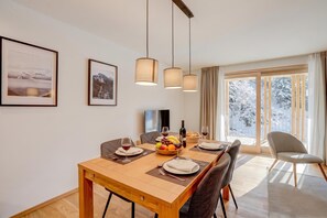 A dining area with a wooden table set for two, a television, and a view of a snowy landscape through a large window. - Residenza Castanea 306 San Bernardino Alpine Apartments, San Bernardino, Switzerland (San Bernardino)
