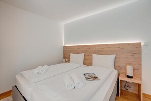 A double bed with white linens and rolled towels, a wooden headboard, a bedside table with a lamp, and a magazine on the bed. - Residenza Castanea 306 San Bernardino Alpine Apartments, San Bernardino, Switzerland (San Bernardino)