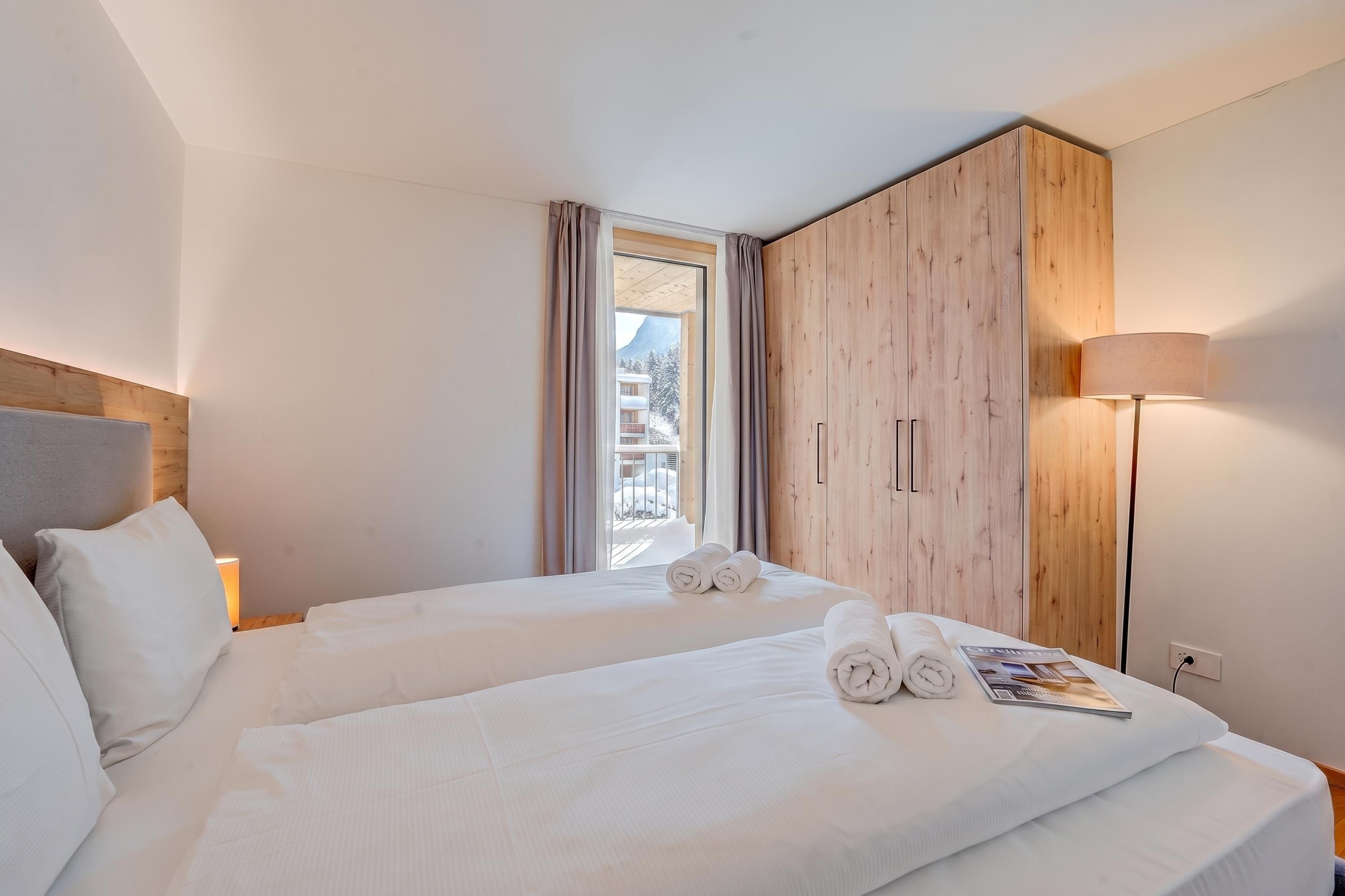 Une chambre d’hôtel avec deux lits, une armoire en bois, une fenêtre avec des rideaux, une lampe de chevet et un magazine sur le lit.