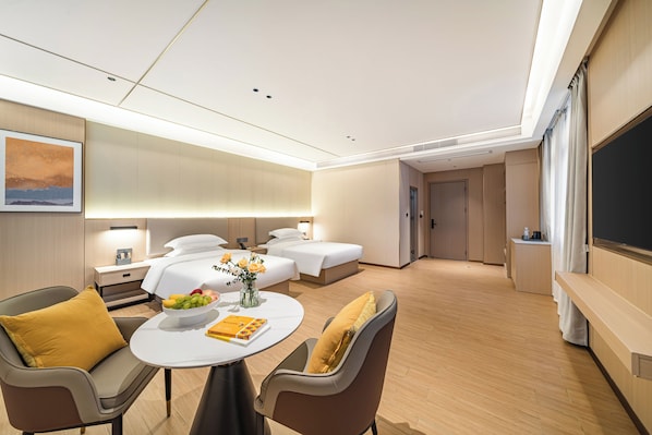 A modern hotel room with a bed, two bedside tables, a seating area with a table, and a large flat-screen TV.