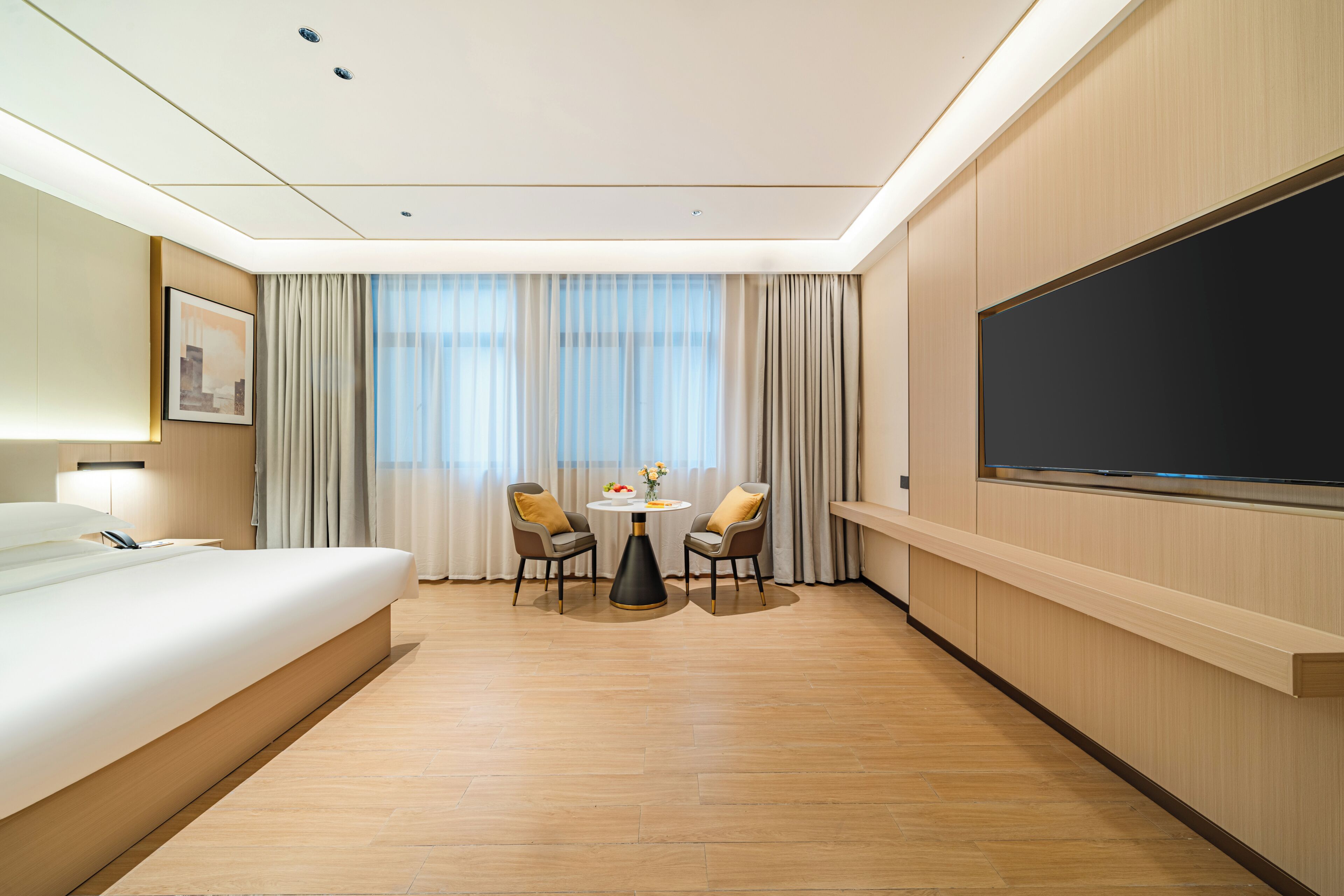 Habitación de hotel con cama, escritorio, dos sillas, televisión y un ventanal con cortinas.