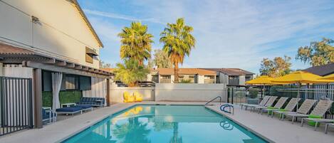A swimming pool with lounge chairs and umbrellas, surrounded by buildings and palm trees.