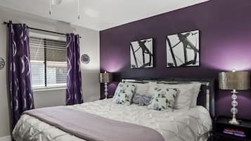 Une chambre à coucher avec un lit, deux lampes de chevet, une fenêtre avec des rideaux violets et deux œuvres d’art murales.