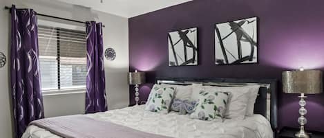 A bedroom with a bed, two bedside lamps, a window with purple curtains, and two wall art pieces.