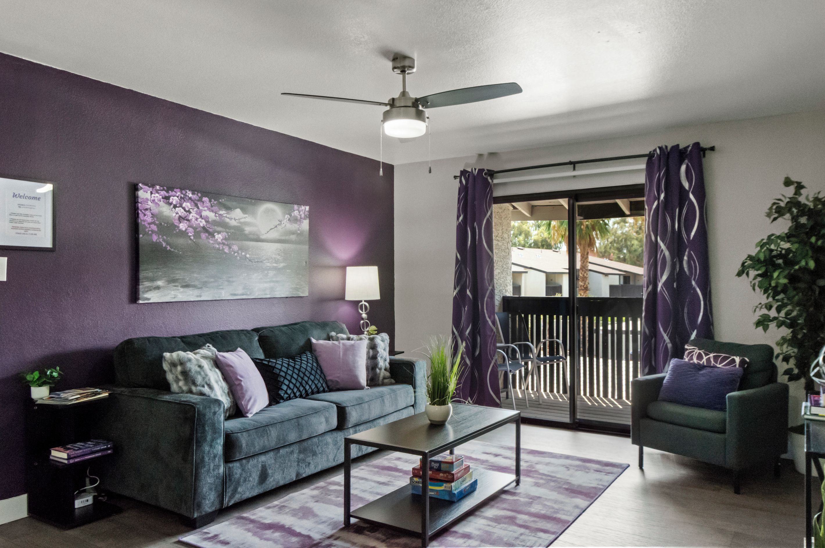 A living room with a sofa, a coffee table, and a chair. There is a ceiling fan, a painting on the wall, and a window with curtains.