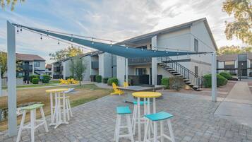 Un espace extérieur aménagé en patio, doté d’un coin salon couvert, d’un escalier menant à un niveau supérieur et offrant une vue sur les bâtiments adjacents.