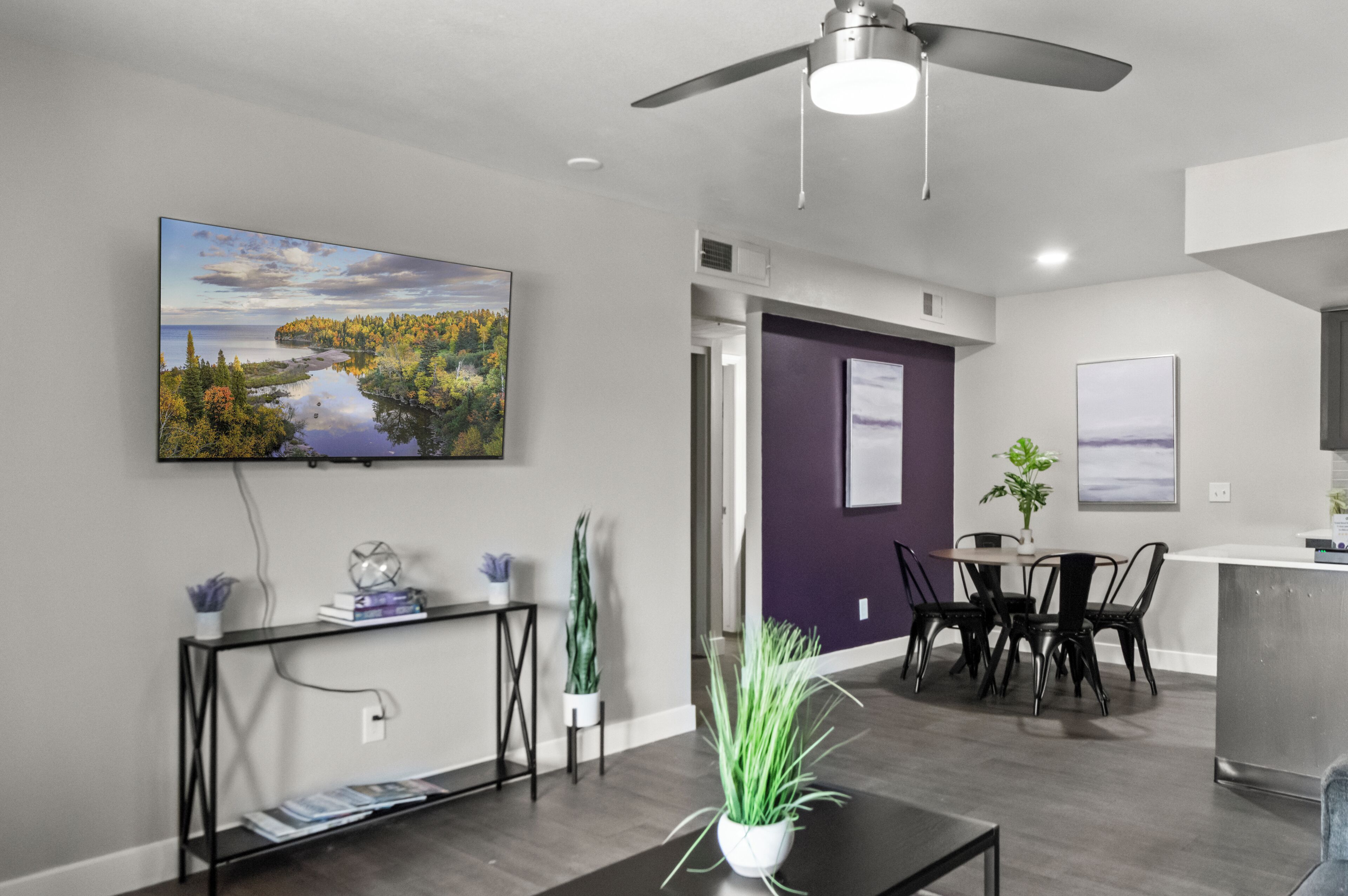 A modern living room with a flat-screen TV mounted on the wall, a dining area with a table and chairs, and a kitchen area with a counter and cabinets.