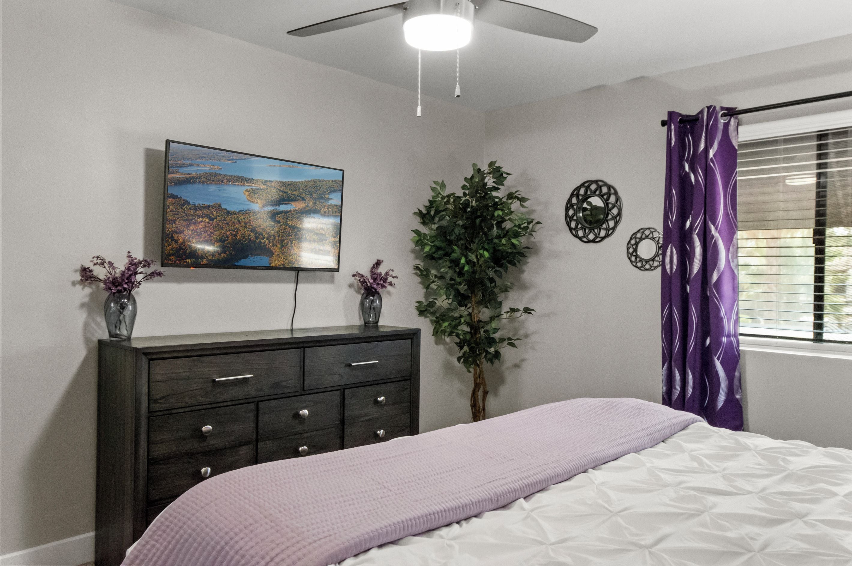 A bedroom with a bed, dresser, TV, plant, and window with curtains.