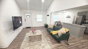 A living room with a green sofa, a coffee table with flowers, a TV, and a kitchen area with a refrigerator. - Enjoyable 4-bedroom house with AC in charming Atlanta (Atlanta)