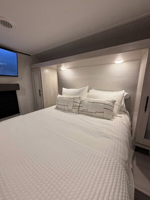 A bedroom with a large bed, a headboard with built-in lighting, a television mounted on the wall, and a wardrobe. - Prairie Moon Charming 1-bedroom recreational vehicle with AC in Springtown (Springtown)