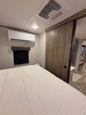 A hotel room with a bed, an air conditioning unit, a wooden wardrobe, and a bathroom with a toilet and sink. - Prairie Moon Charming 1-bedroom recreational vehicle with AC in Springtown (Springtown)