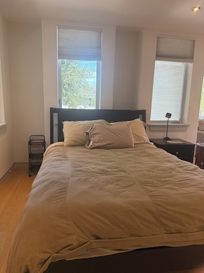 Un dormitorio con una cama, dos ventanas con persianas, una mesita de noche y una lámpara.
