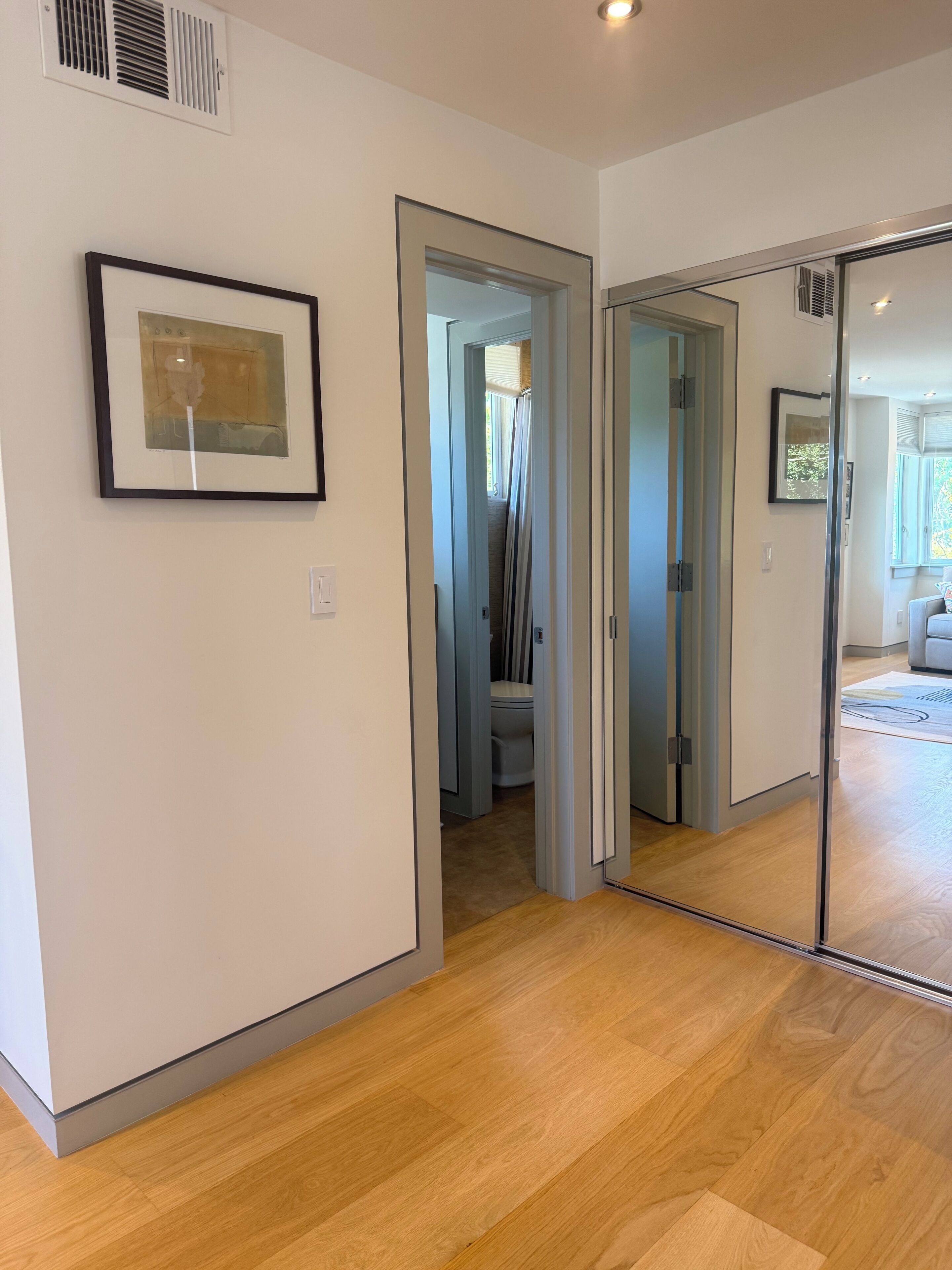 Een gang met een kunstwerk in een lijst, een deur naar een badkamer en een spiegel die een kamer met een raam weerspiegelt.