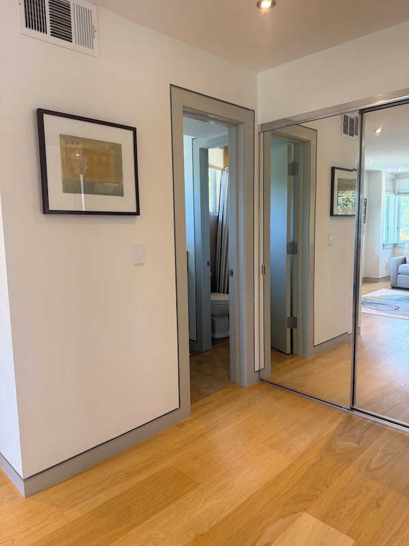 Un pasillo con una obra de arte enmarcada, una puerta que da al baño y un espejo que refleja una habitación con ventana.
