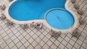 Ein großer, nierenförmiger Pool mit einem kleineren Abschnitt, umgeben von Liegestühlen und Tischen.