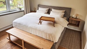 A neatly made bed with a wooden bench, a bedside table with a lamp, and a view of greenery outside. - The OAK SUITE by Interhome (Bad Bederkesa)