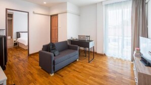 Una habitación de hotel moderna con piso de madera, un sofá gris, un escritorio con una computadora portátil, un televisor y un amplio ventanal con cortinas.