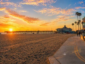 A beach with a pier extending into the water, a building with a dome roof, and a concrete walkway with palm trees. - Coastal Chic Retreat | Steps to the Sand (Newport Beach)