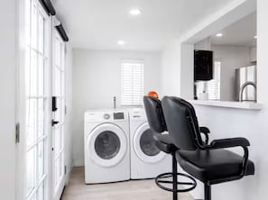 A laundry area with a washing machine and dryer, a chair, and a mirror above a countertop. - Coastal Chic Retreat | Steps to the Sand (Newport Beach)