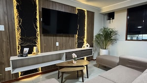 Ein modernes Wohnzimmer mit einem großen Flachbildfernseher, der an einer Wand mit einem besonderen strukturierten Design angebracht ist. Es gibt eine Sofagarnitur und einen kleinen runden Tisch mit einer Vase.
