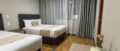 A hotel room with two beds, a desk, a chair, a small table with a card, and a potted plant.