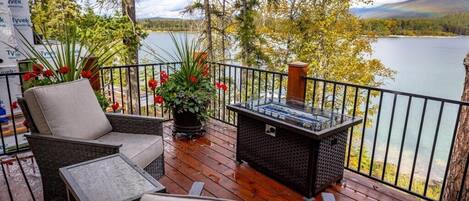 A wooden deck with outdoor furniture, a fire feature, and a view of a lake and forest.