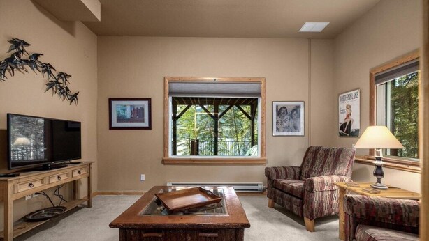A living room with a television, a fireplace, a window with a view of trees, and a patterned armchair. - Whitefish Lake Waterfront Retreat W/ Private Dock (Whitefish)
