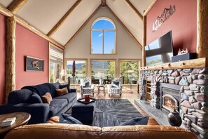 A spacious living room with a fireplace, a large window, and a television mounted on the wall. - Whitefish Lake Waterfront Retreat W/ Private Dock (Whitefish)