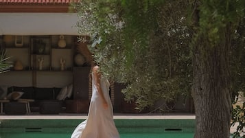A person in a white dress sitting on a bench by a poolside, with a building and trees in the background.