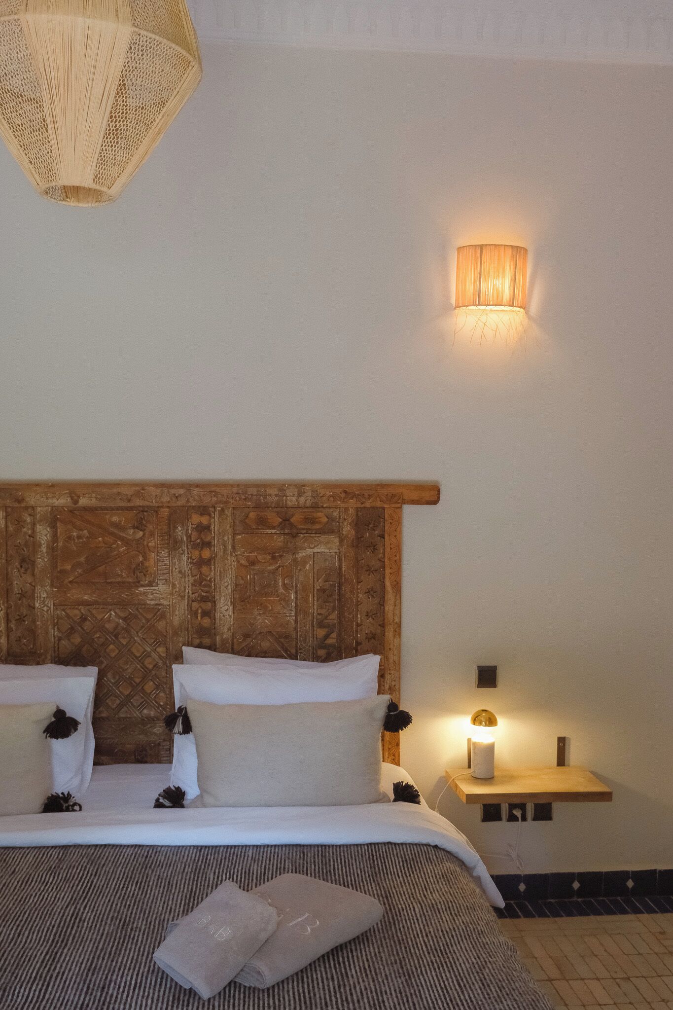A hotel room with a wooden headboard, a bed with white linens, a bedside table with a lamp, and a wall-mounted light fixture.