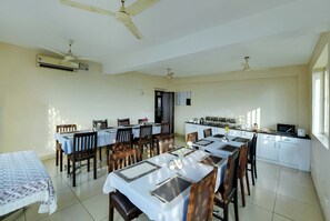 A dining area with tables set for four, ceiling fans, an air conditioning unit, and a kitchen area with appliances. - Avm Comfort Inn (Bengaluru)