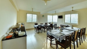 A dining area with multiple tables and chairs, a buffet counter with food items, and ceiling fans. - Avm Comfort Inn (Bengaluru)