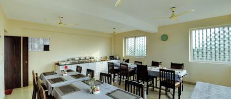 A dining area with tables set for four, ceiling fans, a kitchen area with appliances, and a window with patterned curtains.