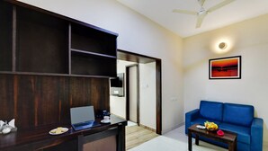 A modern living room with a blue sofa, a wooden desk with a laptop, and a dining table with fruit. - Avm Comfort Inn (Bengaluru)