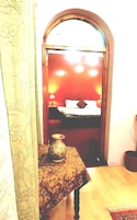 A room with a bed, a small table with a vase, and a mirror reflecting a red wall.