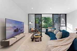 A modern living room with a flat-screen TV, a blue sofa, a coffee table, and a large window offering a view of greenery outside. - Sunny Townhouse Style Apartment at Bondi (Bondi)