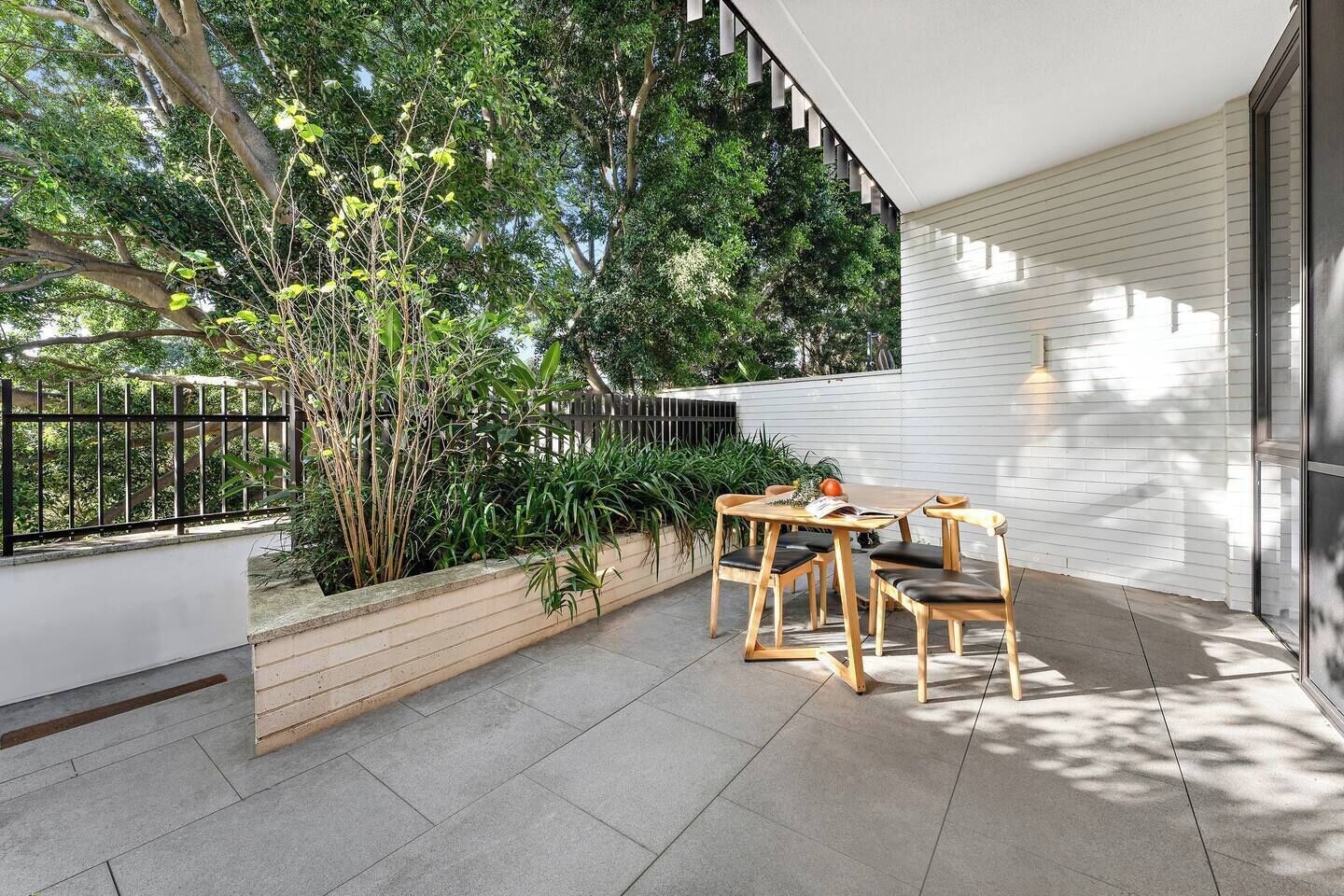 Un patio moderno al aire libre con una mesa y sillas de madera, rodeado de abundante vegetación y un muro blanco.
