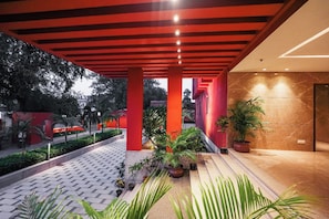 A covered outdoor area with a tiled floor, red structural columns, and potted plants.