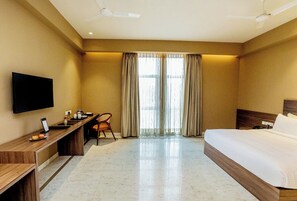 A hotel room with a bed, desk, chair, television, and large windows with curtains. - Gulab Kothi By Grapevine Varanasi (Kashipur)