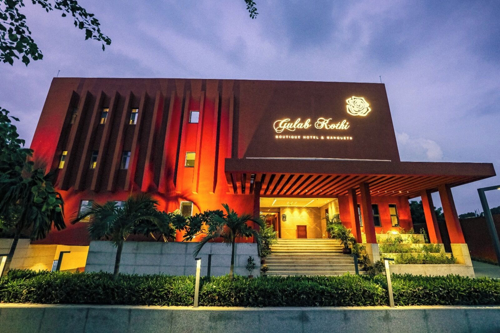 A modern building with a prominent entrance, a sign reading 'Gulab Kothi Boutique Hotel & Banquets,' and a well-lit facade.