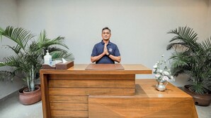 Un mostrador de recepción con un hombre de pie detrás, una computadora portátil sobre la mesa y plantas en macetas a cada lado.