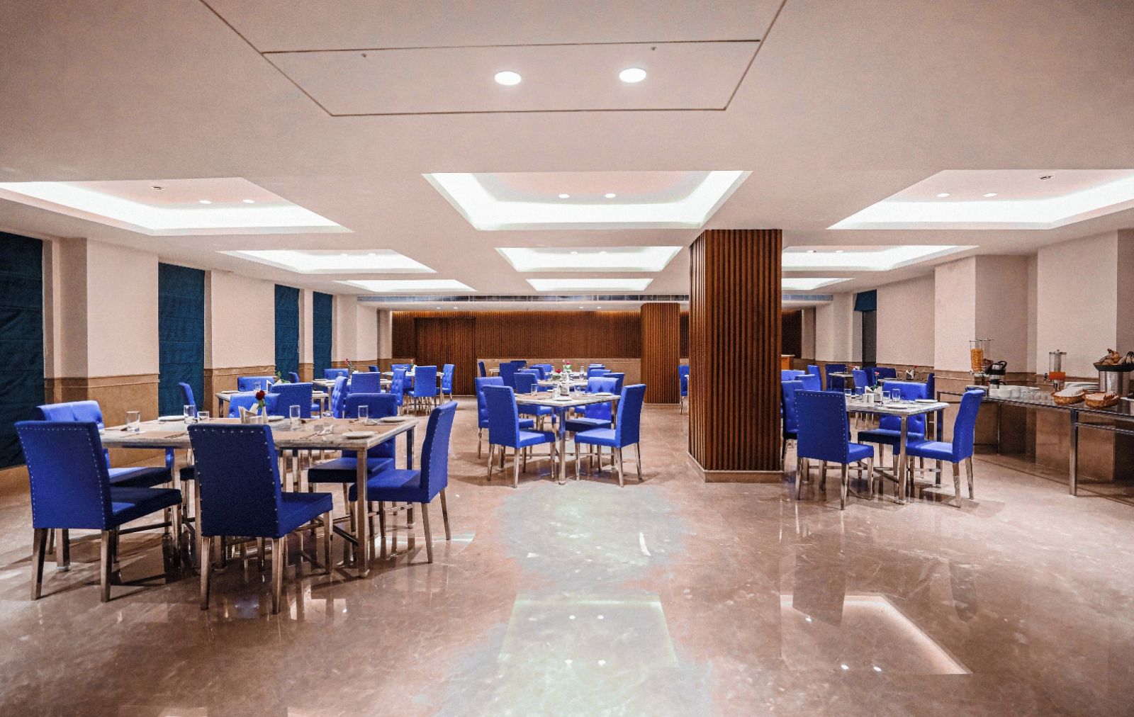 A spacious hotel dining area with multiple tables and chairs, a central column, and a polished floor.