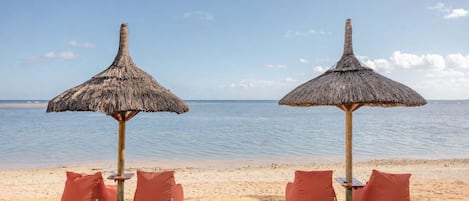 Two thatched beach umbrellas with cushions on a sandy beach, overlooking calm waters.