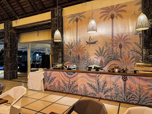 Zona de recepción con un muro decorativo que muestra palmeras y un pescador, un mostrador con una lámpara de fibra y un suelo de baldosas.