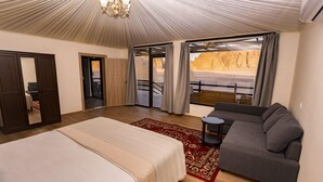 A bedroom with a bed, a sofa, a chandelier, and a view of a desert landscape through large windows. - Sun City Camp (Wadi Rum)