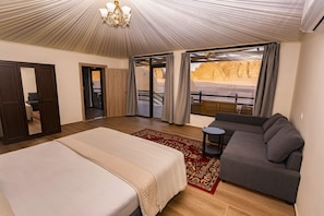 A bedroom with a bed, a sofa, a chandelier, and a view of a desert landscape through large windows. - Sun City Camp (Wadi Rum)